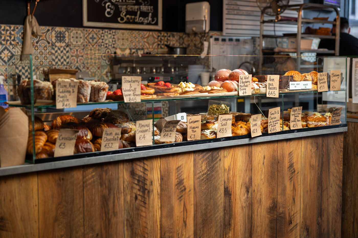 The wood-fronted pastry counter with hand-written tags and patterned tiles behind.